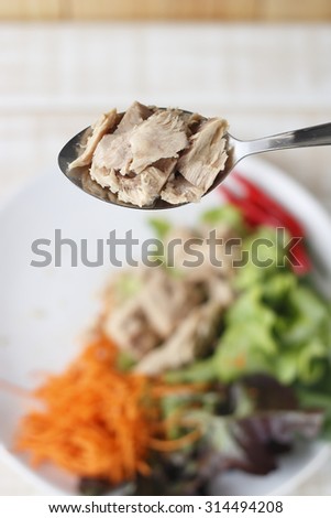 canned tuna inside holding spoon above blur background of tuna and vegetable dish.