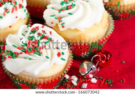 Holiday cupcakes with vanilla frosting and red and green sprinkles. Red holiday background with Christmas bells. Closeup with shallow depth of field.
