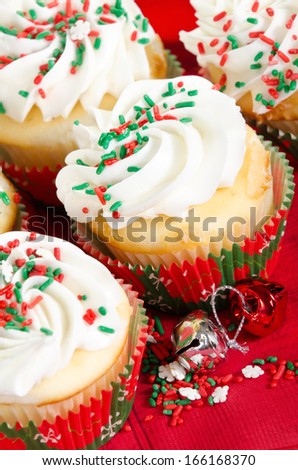Holiday cupcakes with vanilla frosting and red and green sprinkles. Red holiday background with Christmas bells. Closeup with shallow depth of field.