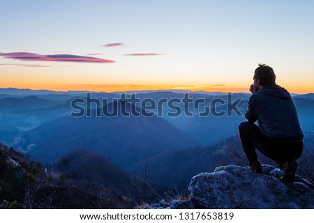 Male hiker crouching on top of the hill and enjoying scenic view of twilight landscape below. Hiking, achievement, expectation, optimism and self-reflection concepts. 商業照片 © 