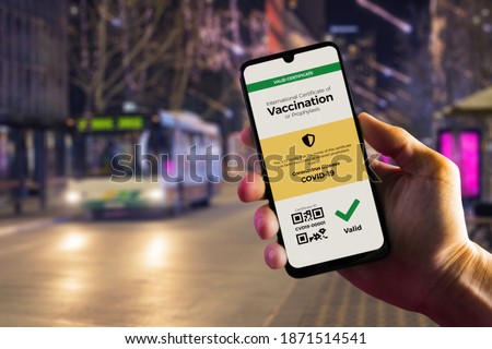 Smartphone displaying a valid digital vaccination certificate for COVID-19 in male's hand, downtown and city bus in background. Vaccination, disease immunity passport, health and surveillance concepts