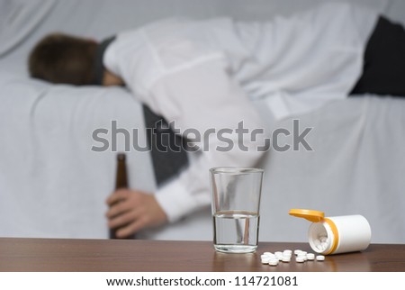 Glass of water and pills on the table. In the background out of focus a passed out businessman on the sofa.