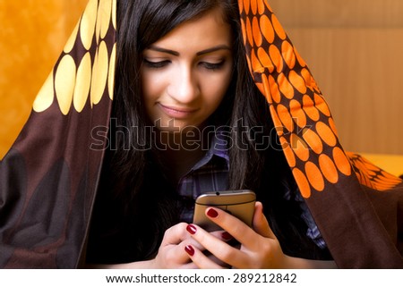 Closeup portrait of beautiful teenage girl using mobil phone hidden under the colorful sheet  in her bedroom