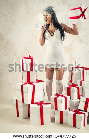 Young woman as angel with heap of gift boxes shouting through megaphone indoors