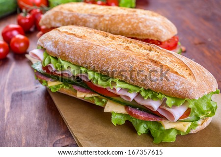 Two Long Ciabatta Sandwiches with lettuce, slices of fresh tomatoes, cucumber, ham, salami and cheese