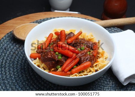 Beef Stew with Carrots & Peas. Serving of beef stew in porcelain bowel made with top round, baby carrots, green peas & served over a bed of egg noodles.