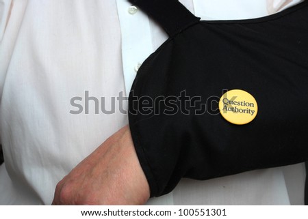 Question Authority Button on Arm Sling. Yellow question authority button pinned onto black arm sling against white button-up shirt.