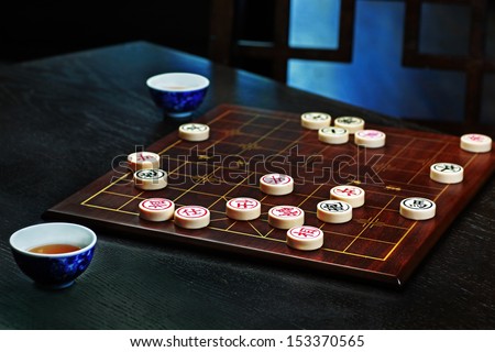 Xiangqi/Chinese Chess Board/Pieces show game in progress on dark wood board with white pieces on dark wood surface & blue design enamel tea cup & chair in background
