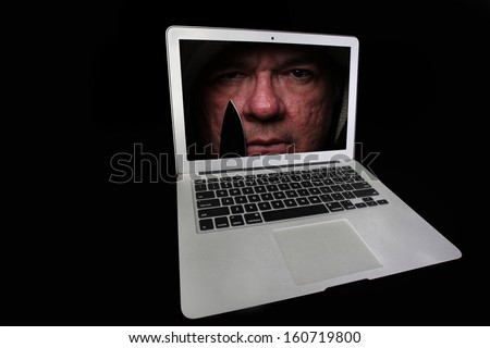 Isolation against black of note book computer with sinister, hooded man holding knife looking out from screen