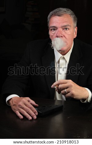 Middle age man seated at table with automatic pistol set before him smoking cigar with smoke coming out of his mouth