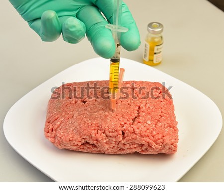 High tech foods series/surgically gloved hand injecting yellow liquid from syringe into raw ground beef with vial of liquid in the background on white platter