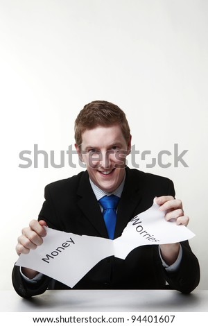 man in a suit sat at a desk ripping a  piece of paper up with the words money worries printed on it and looking happy doing so.