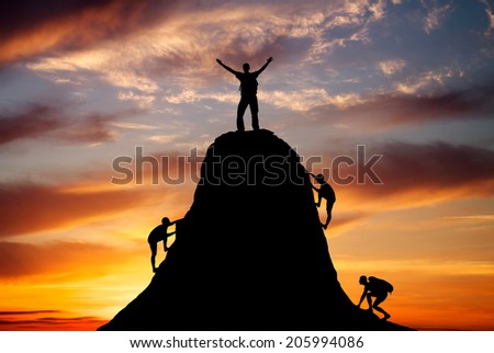 Man on top of the mountain and the other people to climb up on fiery orange background. Rock climber at sunset background. Sport and active life.