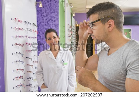 Optical store, people and lenses. Young man looking with glasses and optometris woman in the background.