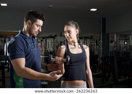 Woman lifting weights at gym training biceps. Personal trainer helps. Focus is in woman.Low key image.