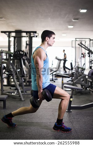 Young man doing legs and biceps exercises with dumbbells at gym with ambient light.