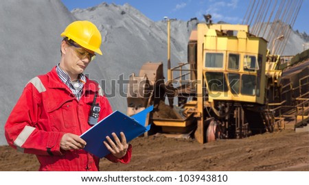 A foreman in a mining site, checking the logs in a blue folder, with a huge wheel digger in the background