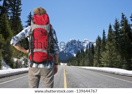 Lonely backpacker looking at the road ahead in beautiful mountain scenics, surrounded by large pine trees and the melting snow in the warm spring sun