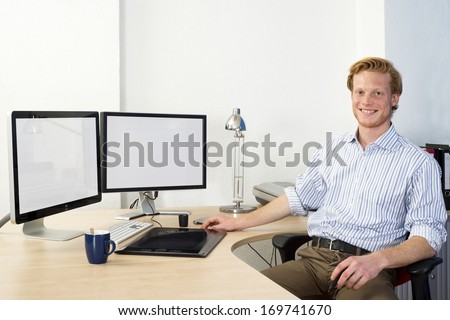 Young Design engineer, using a powerful Computer Aided Design (CAD) workstation sitting confidently behind his desk, smiling
