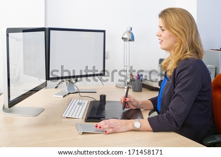 Young woman sitting behind a large desk and a dual screen computer, using a graphic tablet and track pad during design work