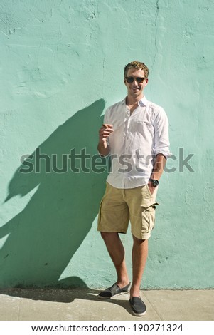 Young man, wearing a white shirt and smoking a cuban cigar, standing in the bright sunlight against a mint green painted, plastered wall