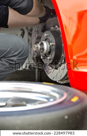Pit crew mechanic working on the brake pads and brake disc of a red race car, with a spare tire in the foreground
