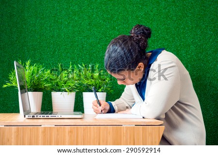Young woman, sitting at a desk with a laptop in front of her, concentrated, writing down notes on a note pad