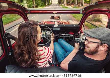 Young couple making a road trip in an old simple comvertibel classic car, looking at eachother and taking pictures.