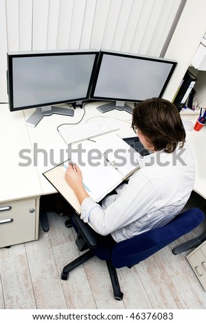 an engineer studying a thick dossier behind his desk with two computer screens on it