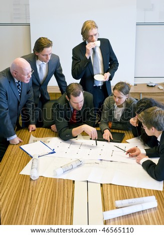 Seven people gathering around the architectural plans, discussing the design in an office cubicle