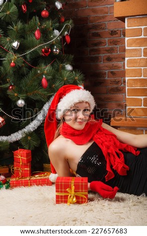 Cute girl in Santa hat and red scarf laying under the Christmas tree with red gift box