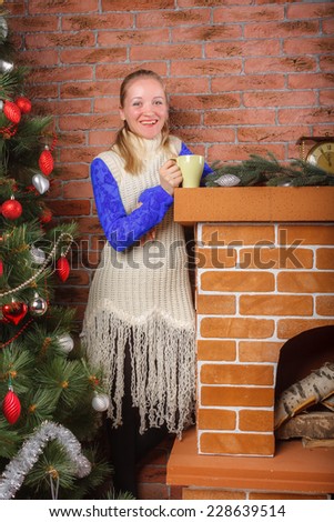 Smiling beautiful woman in a blue lace blouse and white knitted tunic standing with cup of hot drink by the chimney on Christmas