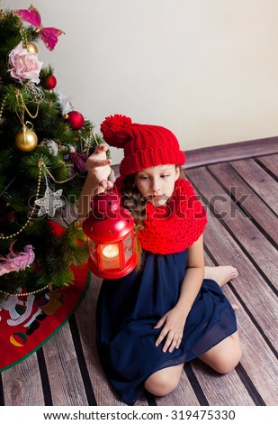 Cute girl in hat and scarf with red lantern sitting by Christmas tree