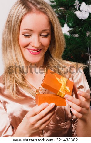 Close up of woman opening small gift box; nice blonde haired woman looking into present box; happy woman with gift nearby Christmas tree; young woman looking surprised while opening gift box