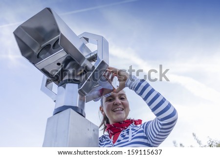 Young smiling woman using coin operated binoculars, against blue sky