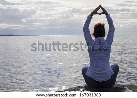 Semi silhouette back light photo-Middle aged  woman in casual clothing  at the seaside meditating and performing Yoga against dramatic cloudy sky