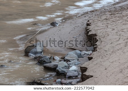 River rocks and washed out river bank next to running river in shaded area/Eroded River Bank with Exposed River Rocks in Shade/Eaten away by river flood is river bank and exposed river rocks