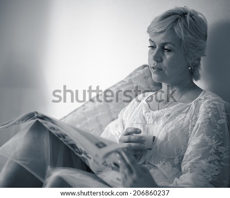 The morning, mature woman reads the newspaper in the bed and drinks juice