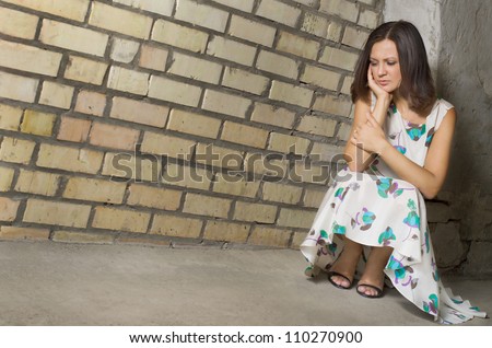 Depressed woman seeking solitude crouched low against a brick wall with her chin on her hand and downcast eyes