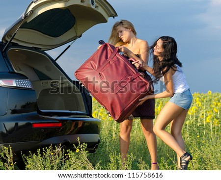 Two women tourists loading a heavy bag into the back of an estate car with the boot open near a field of sunflowers