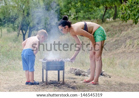 Young boy helping at the barbecue standing fanning the fire with his hat while his mother turns the meat kebabs that are grilling over the coals