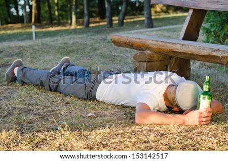 Drunk man sleeping it off in a park lying on his stomach on the ground holding onto his bottle of alcohol with his face turned away from the camera