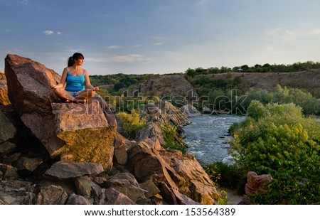 Attractive middle-aged woman sitting cross-legged meditating on a rock above a mountain valley and river as she enjoys the tranquillity of nature