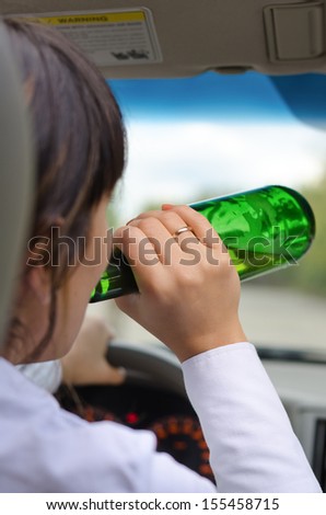 Close up view from behind of an alcoholic woman driver drinking while driving on a road with the bottle of alcohol upended to her lips