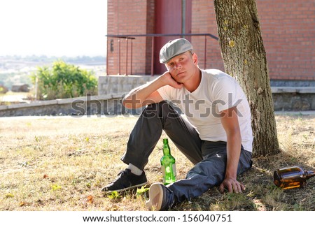 Alcoholic sitting thinking on the grass with his back to a tree surrounded by empty and half full bottles of alcohol