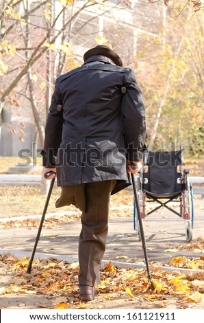 View from behind of a handicapped one-legged man walking on crutches in an autumn park as he heads for his wheelchair which is parked in the street