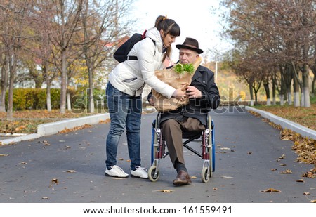 Senior man confined to a wheelchair by a leg amputation being helped with groceries by a woman carer in the street as they return from doing his shopping