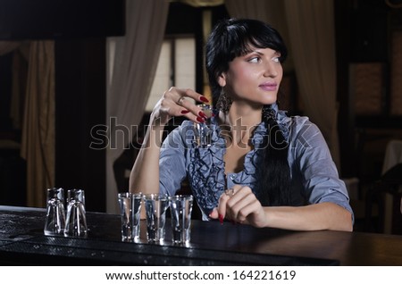 Stylish young woman drinking alone at the bar with a row of full shot glasses lined up on the counter in front of her, looking away to the side