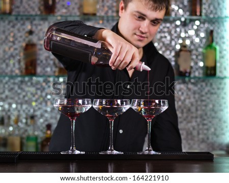 Handsome young male barman mixing cocktails at the bar standing pouring into a row of three glasses