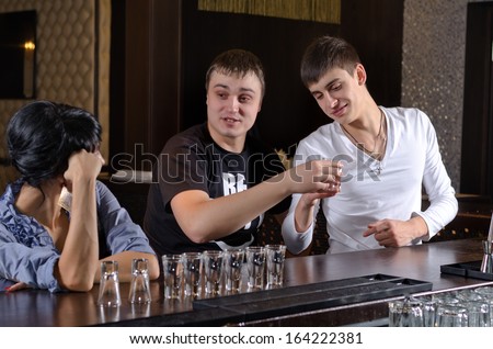 Two attractive young men chatting up a woman at the bar as she sits at the bar counter with a long line of glasses of alcohol in front of her
